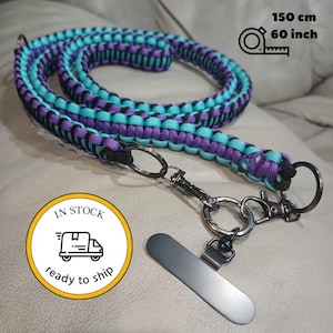 May include: A braided paracord lanyard featuring a repeating pattern of purple, black, and turquoise cords. The lanyard measures 60 inches in length and includes a dark metal swivel lobster clasp, a circular ring, and a flat rectangular metal attachment.