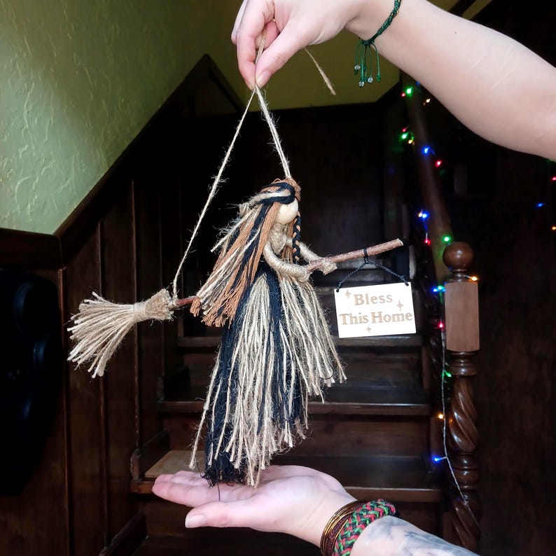 May include: A handmade witch doll with a broom, crafted from natural twine and fabric. The doll has long, flowing hair in shades of brown, black, and tan. A small wooden sign reads "Bless This Home". The doll is suspended by twine.