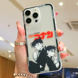 Peut inclure: Coque de téléphone transparente avec un design Détective Conan. Le boîtier a une bordure noire avec le texte "DETECTIVE CONAN" répété. Le design comprend deux personnages de dessins animés et du texte japonais en rouge et orange.