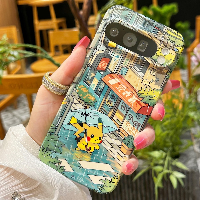 May include: A phone case featuring a colorful illustration of a street scene with a yellow Pikachu character holding an umbrella in the rain. The case has a glossy finish and a detailed design with buildings and shop signs.