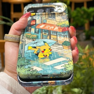 May include: A phone case featuring a colorful illustration of Pikachu holding an umbrella in a rainy street scene. The case has a glossy finish and a silver frame. The design includes a shop with Japanese text and a traffic light.