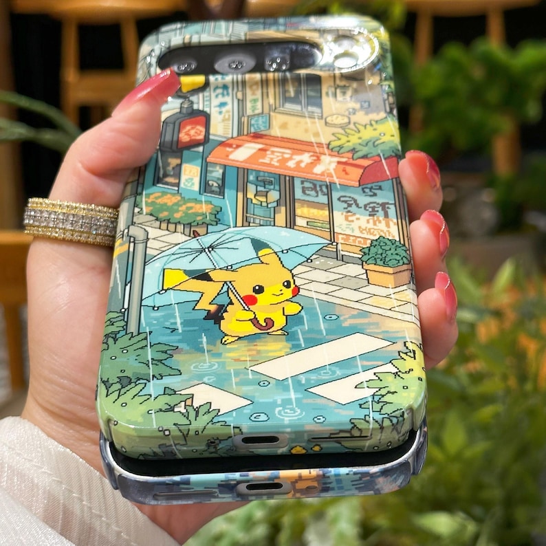 May include: A phone case featuring a colorful illustration of Pikachu holding an umbrella in a rainy street scene. The case has a glossy finish and a silver frame. The design includes a shop with Japanese text and a traffic light.