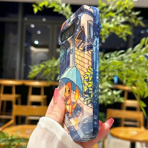 May include: A blue and clear phone case featuring a cartoon illustration of a Charmander character holding a blue umbrella in a rainy street scene. The case has a pixelated art style with a blue and orange color scheme.