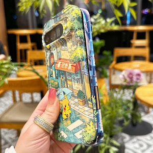 May include: A colorful phone case featuring a detailed illustration of a street scene with buildings, trees, and a cartoon character holding an umbrella. The case has a glossy finish and a protective design.