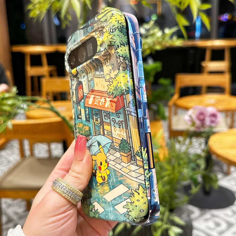 May include: A colorful phone case featuring a detailed illustration of a street scene with buildings, trees, and a cartoon character holding an umbrella. The case has a glossy finish and a protective design.