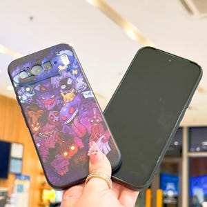 Puede incluir: Una funda negra para smartphone con un dise&ntilde;o colorido de varios personajes de dibujos animados en tonos morados, rojos y azules. La funda se sostiene junto a un smartphone negro.