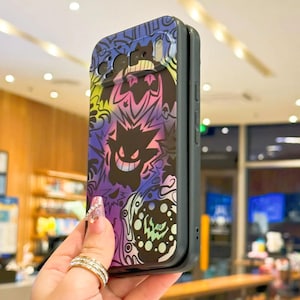 Puede incluir: Una funda de tel&eacute;fono negra y gris con un dise&ntilde;o colorido de personajes de dibujos animados en morado, rosa, amarillo y azul. La funda tiene un acabado brillante y un parachoques protector. El dise&ntilde;o incluye un personaje fantasmal sonriente.