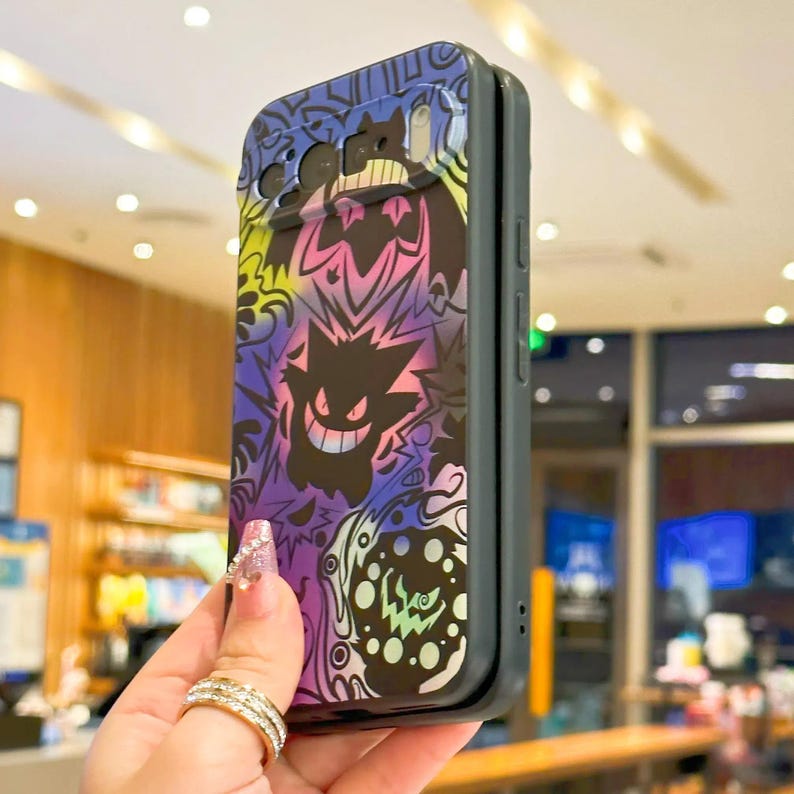 Puede incluir: Una funda de tel&eacute;fono negra y gris con un dise&ntilde;o colorido de personajes de dibujos animados en morado, rosa, amarillo y azul. La funda tiene un acabado brillante y un parachoques protector. El dise&ntilde;o incluye un personaje fantasmal sonriente.