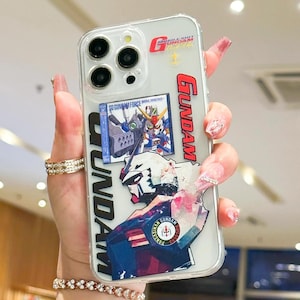 May include: Clear phone case with a Gundam design. The case features multiple Gundam graphics, including the Gundam logo in red, and images of Gundam robots. The case also has the text "MOBILE SUIT GUNDAM" and "SO GUNDAM FORCE".
