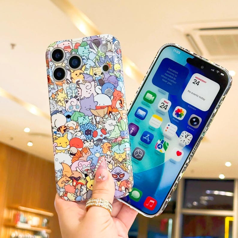 May include: A colorful phone case featuring a pattern of cartoon characters. The case is held next to a smartphone displaying the home screen with app icons and the date, Wednesday 24. The phone case has a clear, glossy finish.