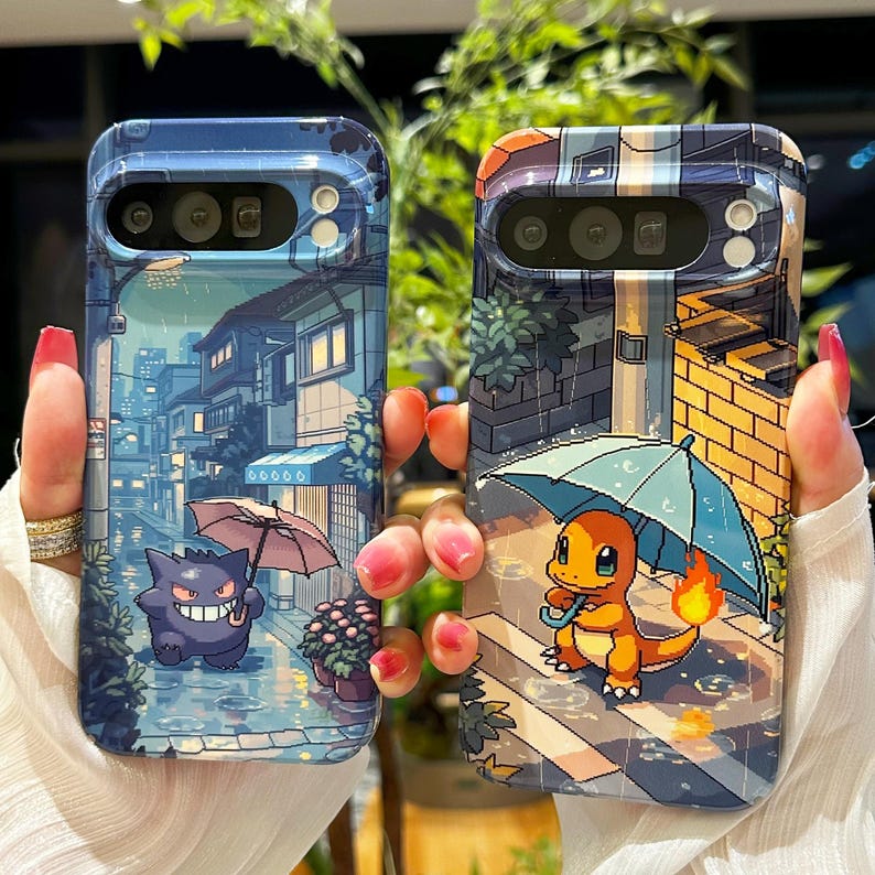 May include: Two phone cases with pixelated illustrations. One case features a purple Gengar with an umbrella in a rainy street scene. The other case shows a Charmander with an umbrella in a similar setting. Both cases have a blue and white color scheme.
