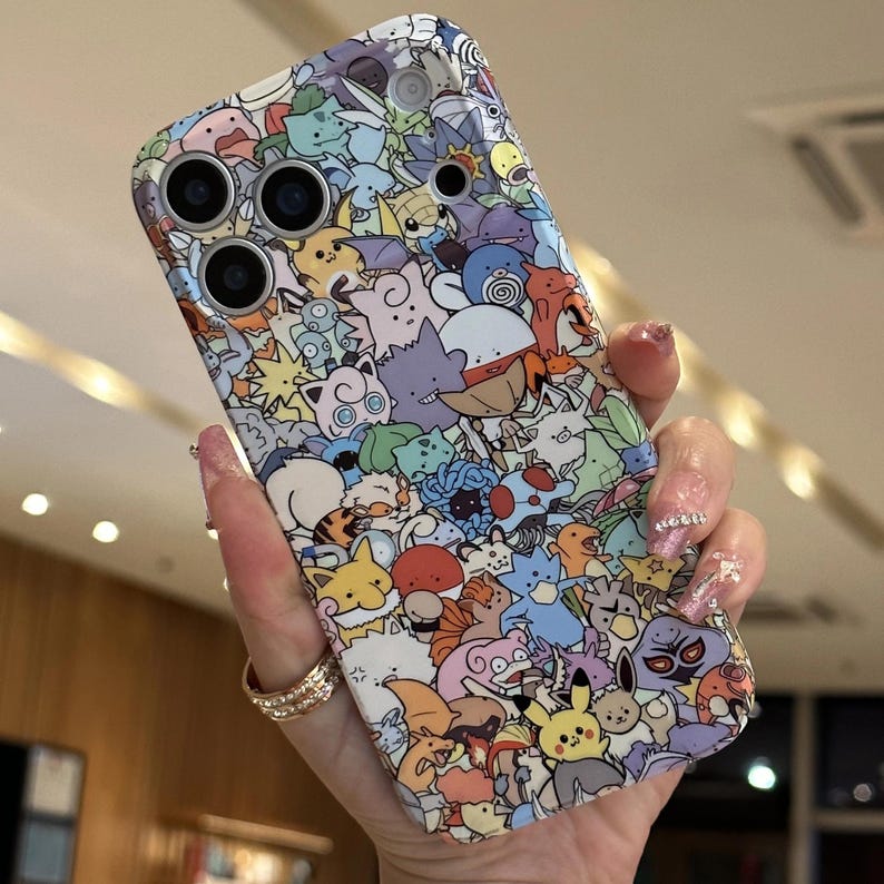 May include: A colorful phone case featuring a repeating pattern of various cartoon characters in a variety of colors. The case has cutouts for the phone's camera lenses and flash. The case is held in a hand.