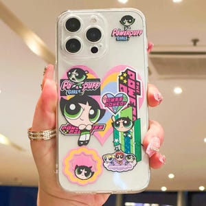 May include: Clear phone case featuring Powerpuff Girls stickers. The stickers include images of the characters Blossom, Bubbles, and Buttercup, along with the words "Powerpuff Girls". The case has a clear background and a glossy finish.