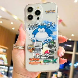 May include: Transparent phone case adorned with a blue Snorlax character and Pokémon stickers. The case has a clear background with the word "Snorlax" in blue and white. Additional stickers include the Pokémon logo and Japanese text.