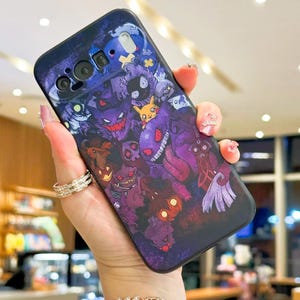 Puede incluir: Funda de tel&eacute;fono azul oscuro con un gr&aacute;fico colorido de varios monstruos de dibujos animados en tonos morados, rojos y amarillos. La funda tiene un acabado brillante y se sostiene en una mano.