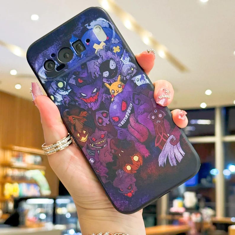 Puede incluir: Funda de tel&eacute;fono azul oscuro con un gr&aacute;fico colorido de varios monstruos de dibujos animados en tonos morados, rojos y amarillos. La funda tiene un acabado brillante y se sostiene en una mano.