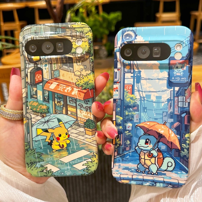 May include: Two phone cases with colorful pixelated illustrations. One case features Pikachu under an umbrella in a rainy street scene. The other case shows Squirtle under an umbrella in a similar setting. Both cases have a camera cutout.