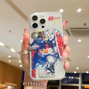 May include: Transparent phone case with a vibrant robot illustration, a red shield, and the word "GUNDAM" in red. The case has a clear border and a design on the back.