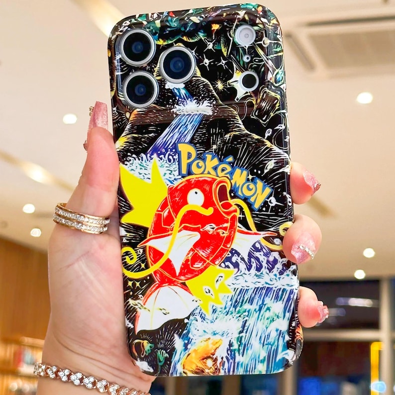 May include: A black and colorful phone case featuring a cartoon image of a red and yellow Magikarp from the Pokemon series. The case has the words "Pokemon" in yellow and blue. The case has a waterfall and mountain design.
