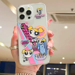 May include: Clear phone case featuring Powerpuff Girls cartoon characters. The case has multiple stickers with the characters, the words "Powerpuff Girls", and other graphic elements in pink, blue, and white. The phone case is held by a person.