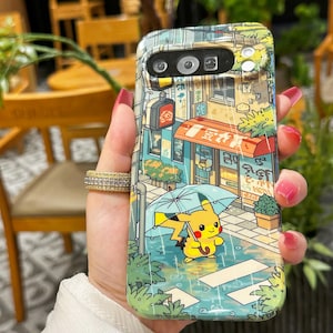 May include: A phone case featuring a colorful illustration of a street scene with a yellow Pikachu character holding an umbrella in the rain. The case has a vibrant design with Japanese text and a red awning.