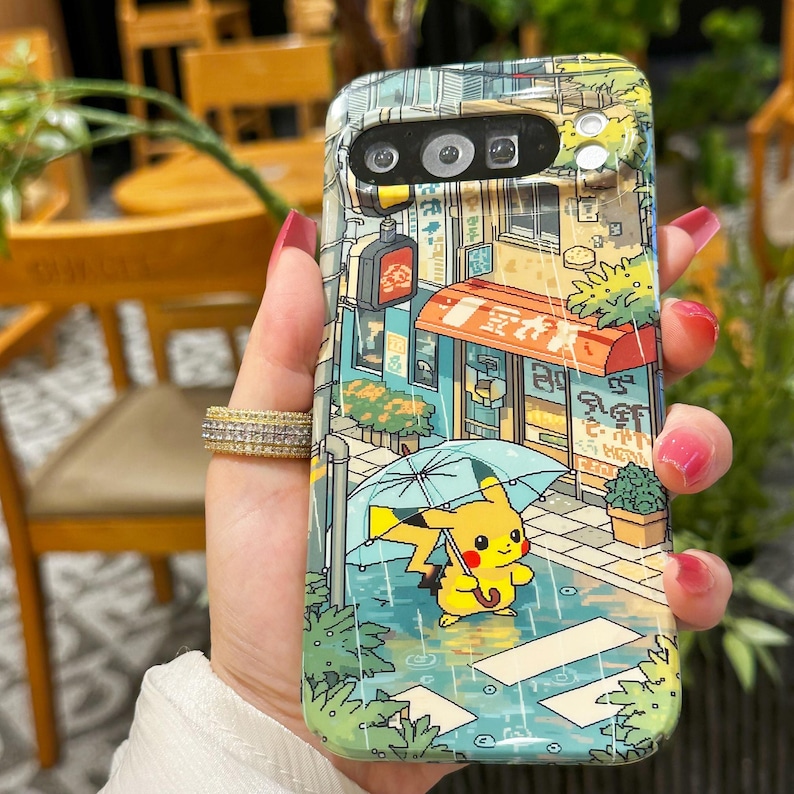 May include: A phone case featuring a colorful illustration of a street scene with a yellow Pikachu character holding an umbrella in the rain. The case has a vibrant design with Japanese text and a red awning.