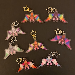 May include: A selection of bat keyrings with vibrant wings and star-shaped attachments. The keyrings display a range of colours, including rainbow, purple, pink, and orange. The bats have detailed faces and are arranged on a dark background.