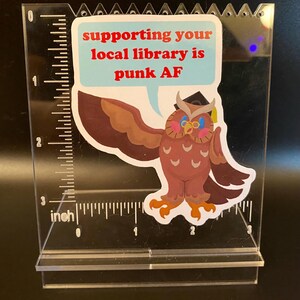 Puede incluir: Un expositor de acrílico transparente con una pegatina que presenta un búho de dibujos animados con un gorro de graduación. El búho tiene gafas azules y habla desde un bocadillo que dice: "apoyar a tu biblioteca local es punk AF."