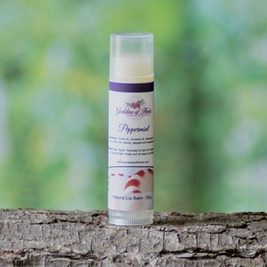 Könnte beinhalten: Ein weißer Stift mit natürlichem Lippenbalsam mit einem violetten Etikett, auf dem "Goddess of Herbs" und "Peppermint" steht. Der Stift liegt auf einem Stück Baumrinde.