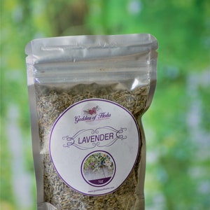 May include: A silver resealable pouch filled with dried lavender flowers. The pouch has a white label with the text "Goddess of Herbs" and "LAVENDER" in purple lettering. The label also includes the text "1 oz" and a website address.