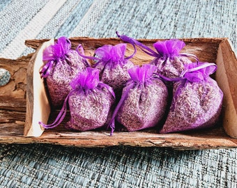 May include: Six small purple fabric pouches filled with dried lavender flowers are arranged in a wooden bowl. The pouches are tied with purple ribbon and have a delicate, sheer texture.
