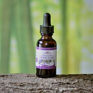 May include: A brown glass bottle with a dropper top, labeled "Goddess of Herbs Herbal Supplement GINKGO BILOBA" in purple text on a white background. The bottle is sitting on a piece of tree bark.