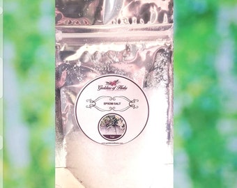 May include: A silver resealable pouch filled with white Epsom salt. The pouch has a label with the text "Goddess of Fleets" and "Epsom Salt" in a pink circle with a floral design.