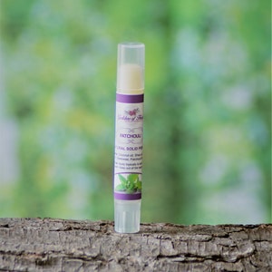 May include: A white tube with a purple label that says "Goddess of Fire" and "Patchouli Natural Solid Perfume". The label also includes a list of ingredients and instructions for use. The tube is sitting on a piece of tree bark.