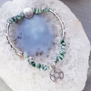 May include: A silver bangle bracelet with a green stone charm and a silver pentagram charm. The bracelet is on a white agate slice.
