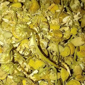 May include: Close-up of dried chamomile flowers, showcasing a mix of yellow and white petals and green stems. The image highlights the natural texture and detail of the herbal tea ingredient, perfect for tea enthusiasts.