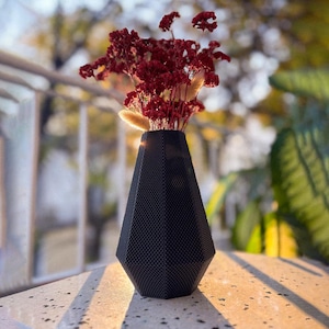 Può includere: Un vaso nero strutturato con un design geometrico, che contiene un bouquet di fiori rossi secchi ed erbe beige. Il vaso ha una base larga e si assottiglia verso l'alto. La composizione è su una superficie macchiata.