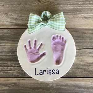 Op de afbeelding: Een ronde witte keramische hanger met een groen-witte gingham strik. De hanger heeft een paarse hand- en voetafdruk met de naam "Larissa" in paars.