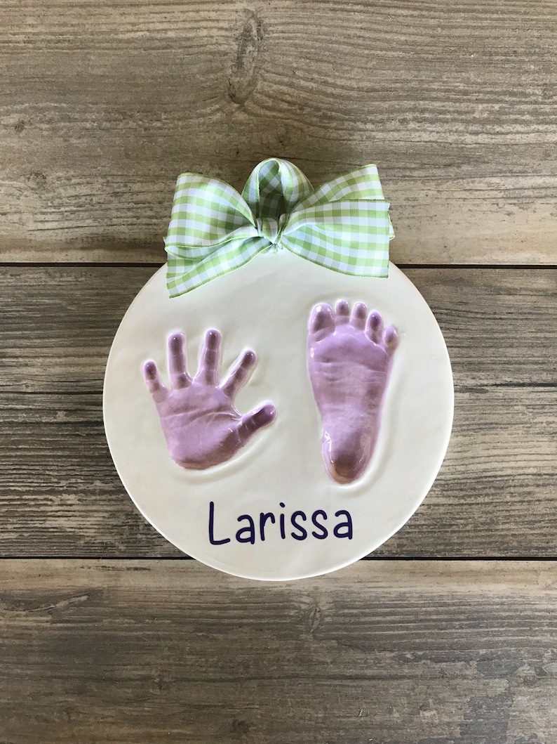 Op de afbeelding: Een ronde witte keramische hanger met een groen-witte gingham strik. De hanger heeft een paarse hand- en voetafdruk met de naam "Larissa" in paars.