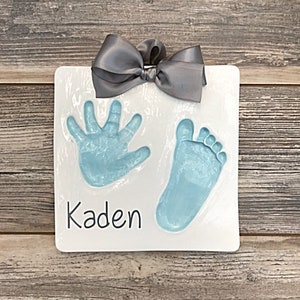 May include: White ceramic plaque with a blue handprint and footprint, a gray bow, and the name "Kaden" written in black.