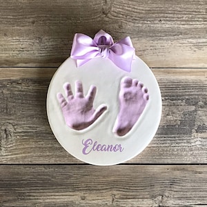 May include: A white ceramic round plaque with a purple ribbon, a handprint and footprint in purple paint, and the name "Eleanor" written in purple.