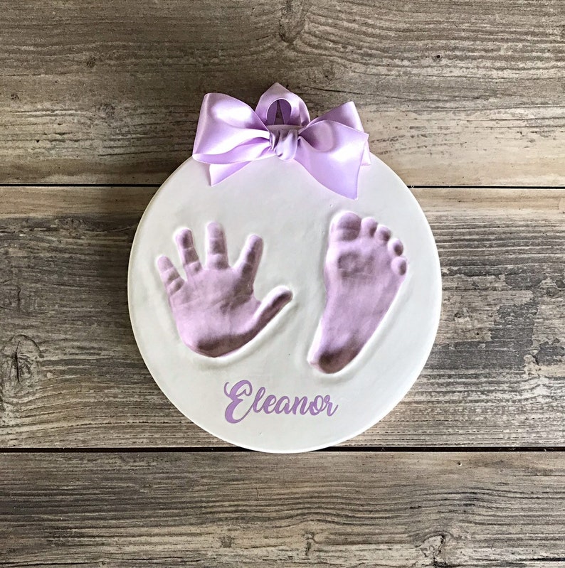 Op de afbeelding: Een ronde witte keramische plaquette met een paars lint, een handafdruk en een voetafdruk in paarse verf, en de naam "Eleanor" in paars.