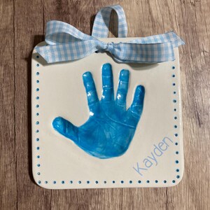 May include: White ceramic handprint plaque with a blue handprint and the name "Kayden" written in blue script. The plaque has a blue and white gingham ribbon attached.