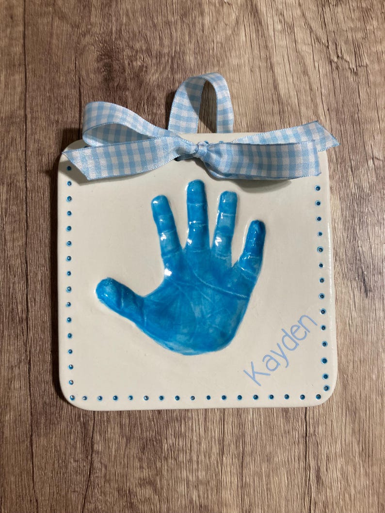 May include: White ceramic handprint plaque with a blue handprint and the name "Kayden" written in blue script. The plaque has a blue and white gingham ribbon attached.