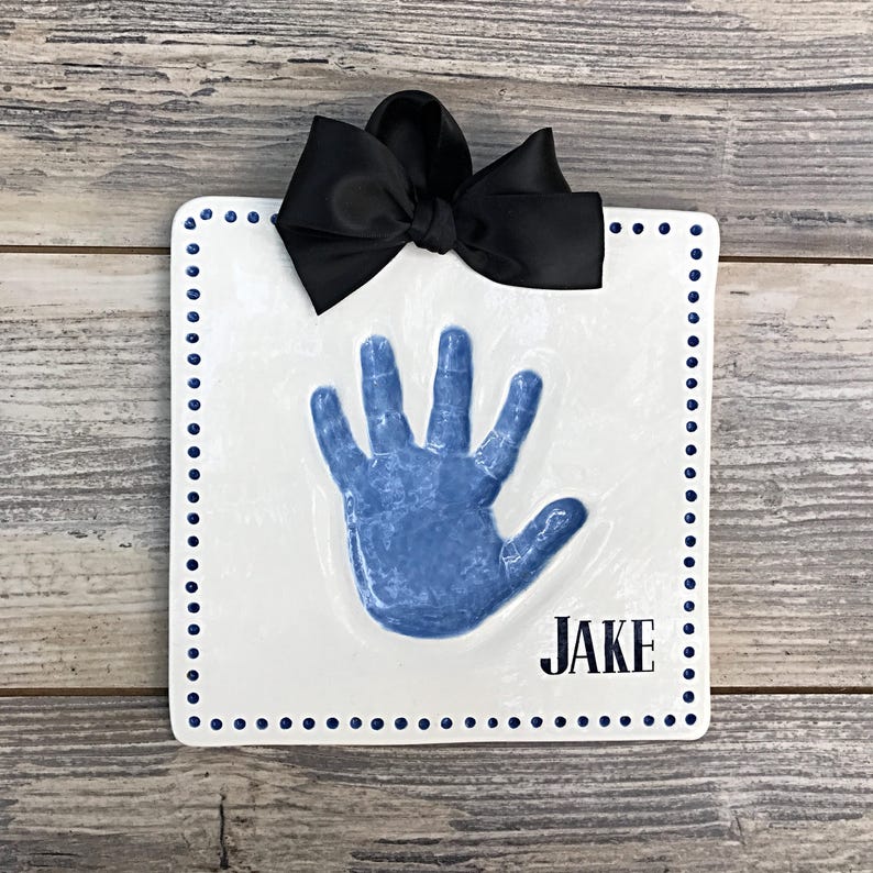 May include: A white ceramic handprint plaque with a blue handprint and the name "JAKE" in black lettering. The plaque has a blue dotted border and a black satin bow.