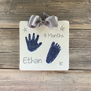 May include: White plaque with a blue handprint and footprint, a silver bow, and the text "4 Months" and "Ethan".