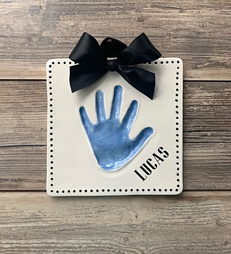 May include: White ceramic handprint plaque with a blue handprint and the name "Lucas" in black lettering. A black bow is attached to the top of the plaque.