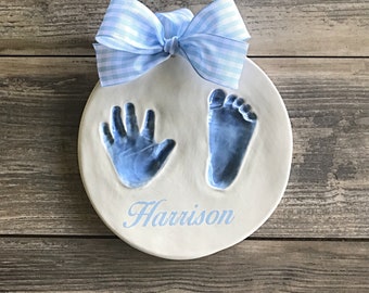 May include: A white ceramic plate with blue hand and footprint impressions. The plate has a blue and white gingham bow on top and the name "Harrison" written in blue cursive.