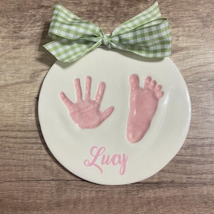 Op de afbeelding: Een wit keramiek bord met roze hand- en voetafdrukken en de naam "Lucy" in roze cursief.