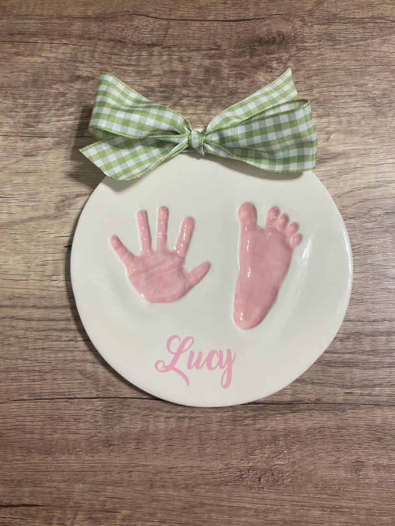 Op de afbeelding: Een wit keramiek bord met roze hand- en voetafdrukken en de naam "Lucy" in roze cursief.